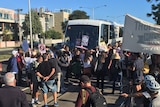 Protesters stand in front of a bus with signs and banners.