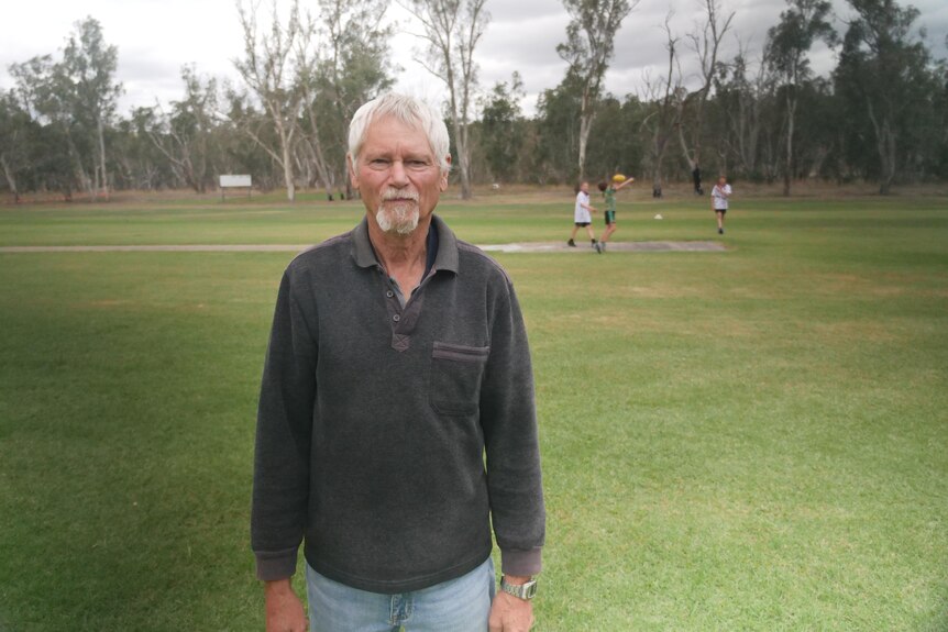 An indigenous man with white hair stands on a cricket pitch. A white tent is visible in the background