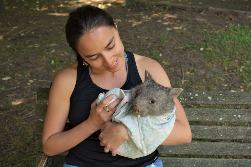 A woman holds a wombat wrapped in cloth, sitting on a bench.