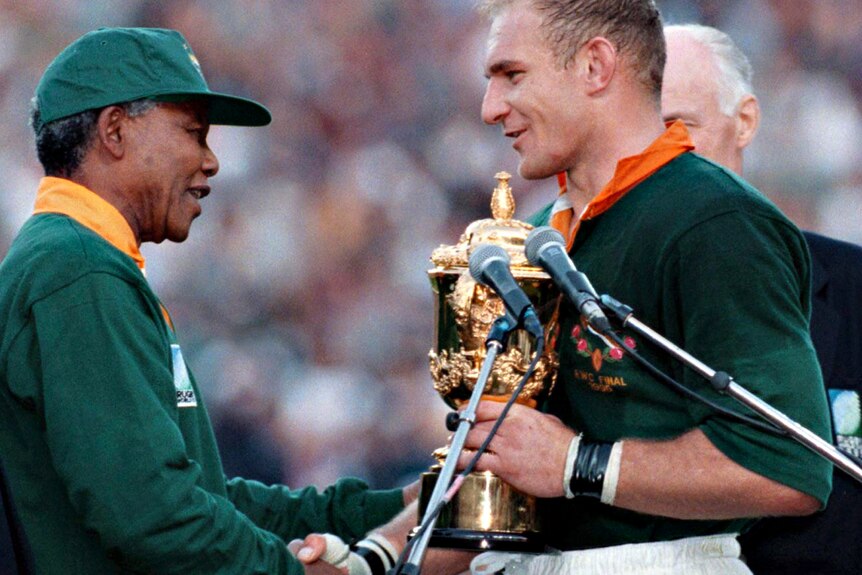 Nelson Mandela, in green jacket and green cap, smiles and shakes hands with Francois Pienaar, in green shirt and white shorts.