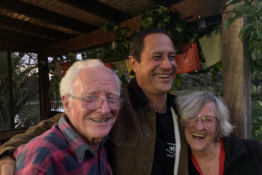 Flood-affected couple smiling as they are hugged by their rescuer.