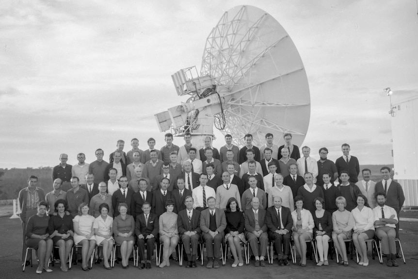 A group of people stand in front of a large satellite dish