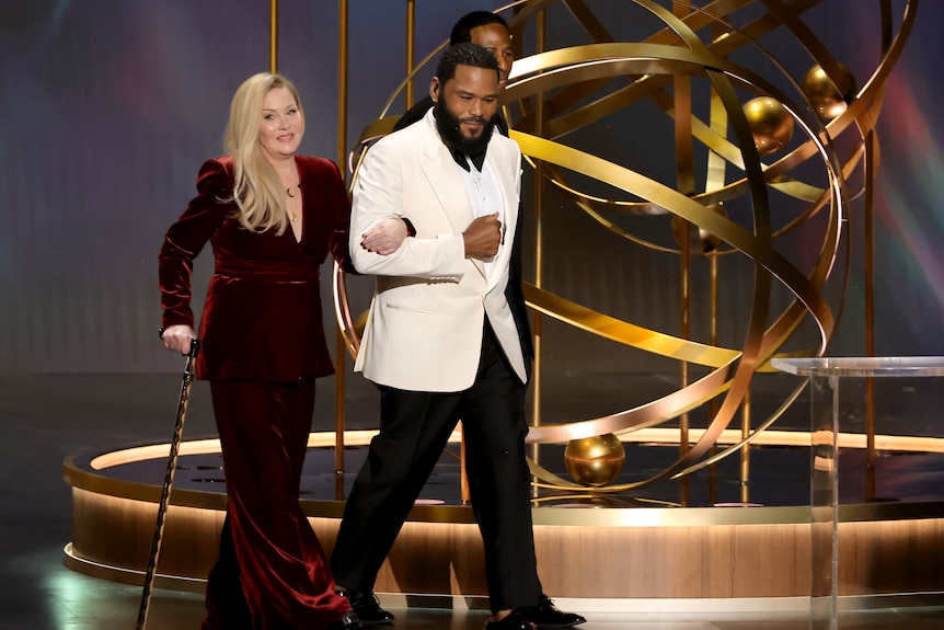 Christina Applegate in a velvet suit holding a cane with other arm being held by Anthony Anderson leading her on stage