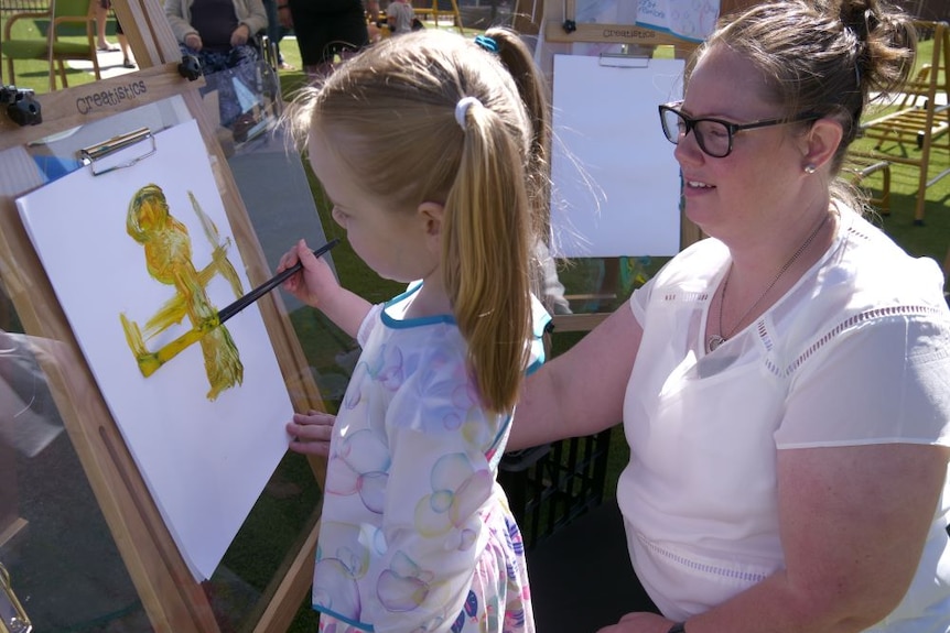 A young girl uses yellow paint on an easel while her mother watches