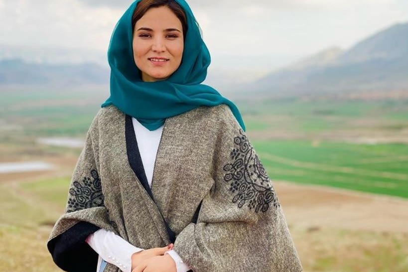 A woman wearing a green hijab and grey shawl smiles against a backdrop of grass and mountains.