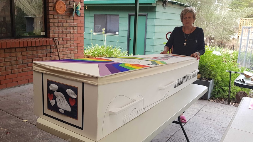 A woman stands behind a long white coffin. The coffin has colourful drawings on it.