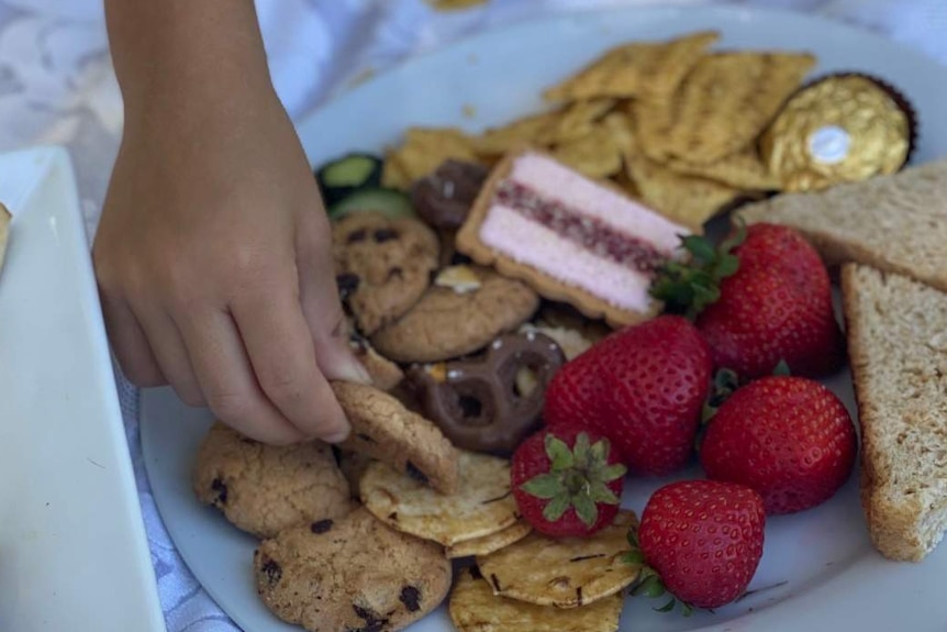 Child's hand picks up a cookie from a plate of food that has strawberries, sandwiches and biscuits