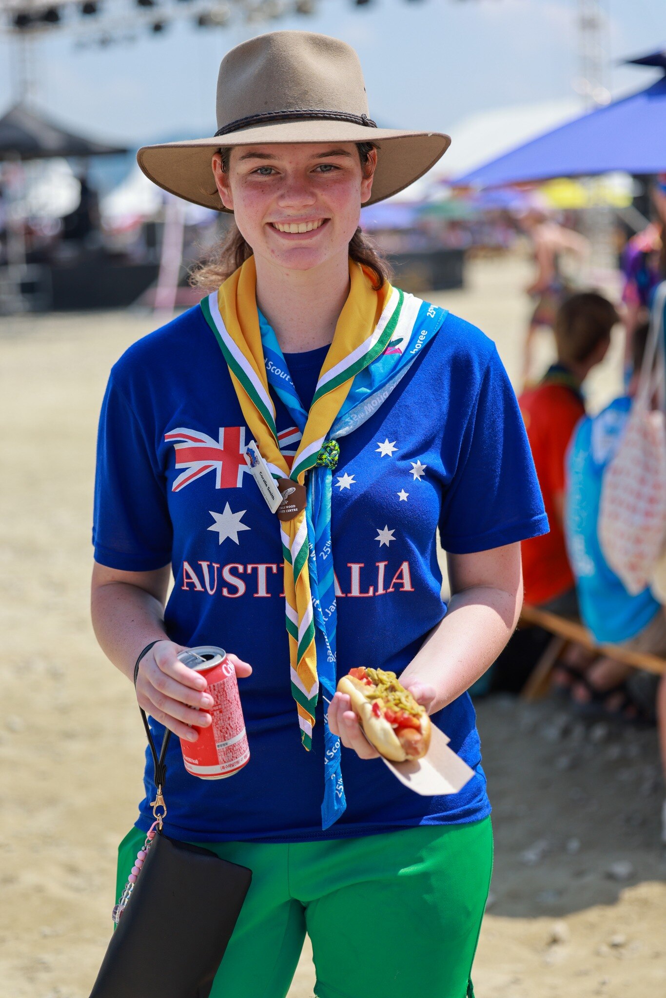 Girl dressed in blue T-shirt with Australia on it, holds sausage in roll and can of drink.