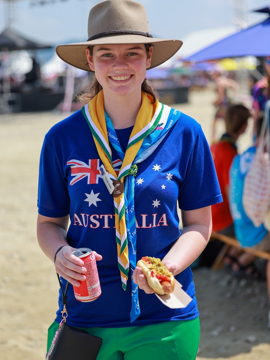 Girl dressed in blue T-shirt with Australia on it, holds sausage in roll and can of drink.