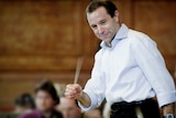 A photo of Mark Wigglesworth in action conducting. He is smiling and holding a baton. An orchestra is out of focus.