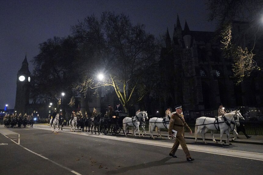 A procession of horses and military members goes down a dark street. Big Ben can be seen in the distance