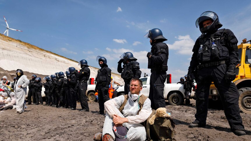 police stand in a row behind a protesters dressed in white sitting on the ground in a mine.