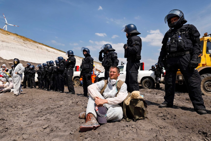 police stand in a row behind a protesters dressed in white sitting on the ground in a mine.