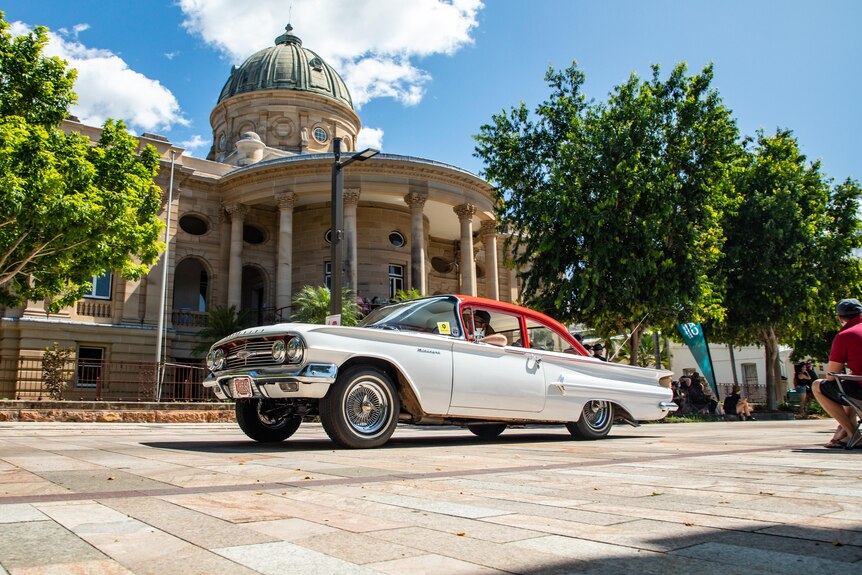 A white and red vintage car drives along a street, with a historic building in the background