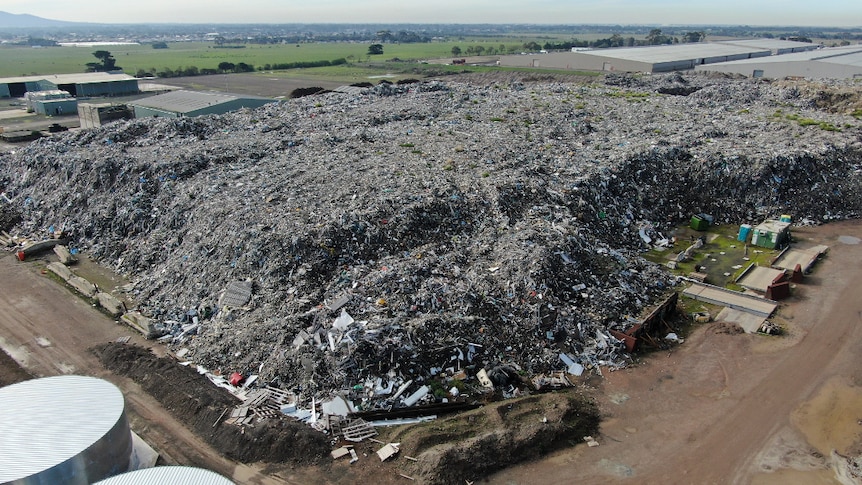 An aerial photo taken by a drone showing a huge pile of waste, bordered by a road, sheds and paddocks. Homes are seen nearby.