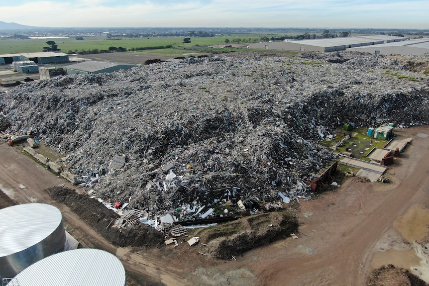 An aerial photo taken by a drone showing a huge pile of waste, bordered by a road, sheds and paddocks. Homes are seen nearby.