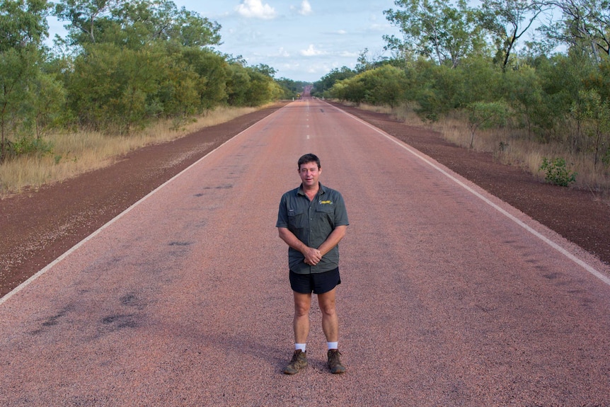 a man standing in the middle of a bitumen road with scrub on either side.