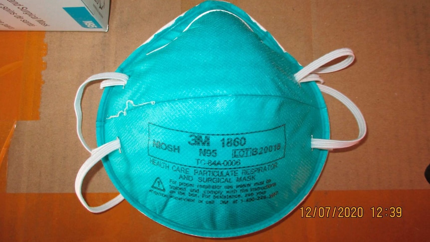 A teal disposable face mask with manufacturing information printed across it in black is seen resting on cardboard.