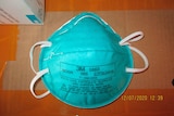 A teal disposable face mask with manufacturing information printed across it in black is seen resting on cardboard.