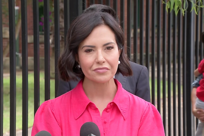 A dark-haired woman in a bright top stands outside and talks to the media.
