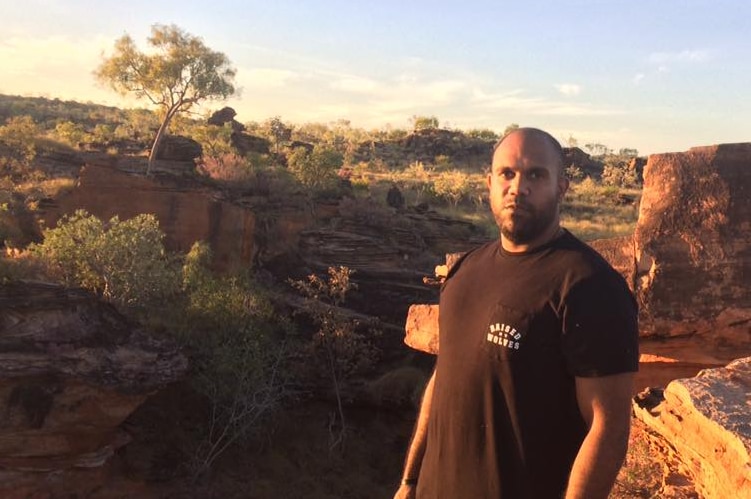 A young man stands in a bush setting wearing a black t-shirt.