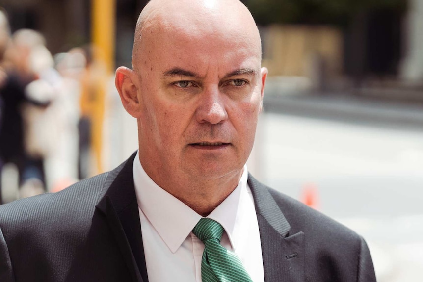 A headshot of a bald man in a suit and green tie outside court.
