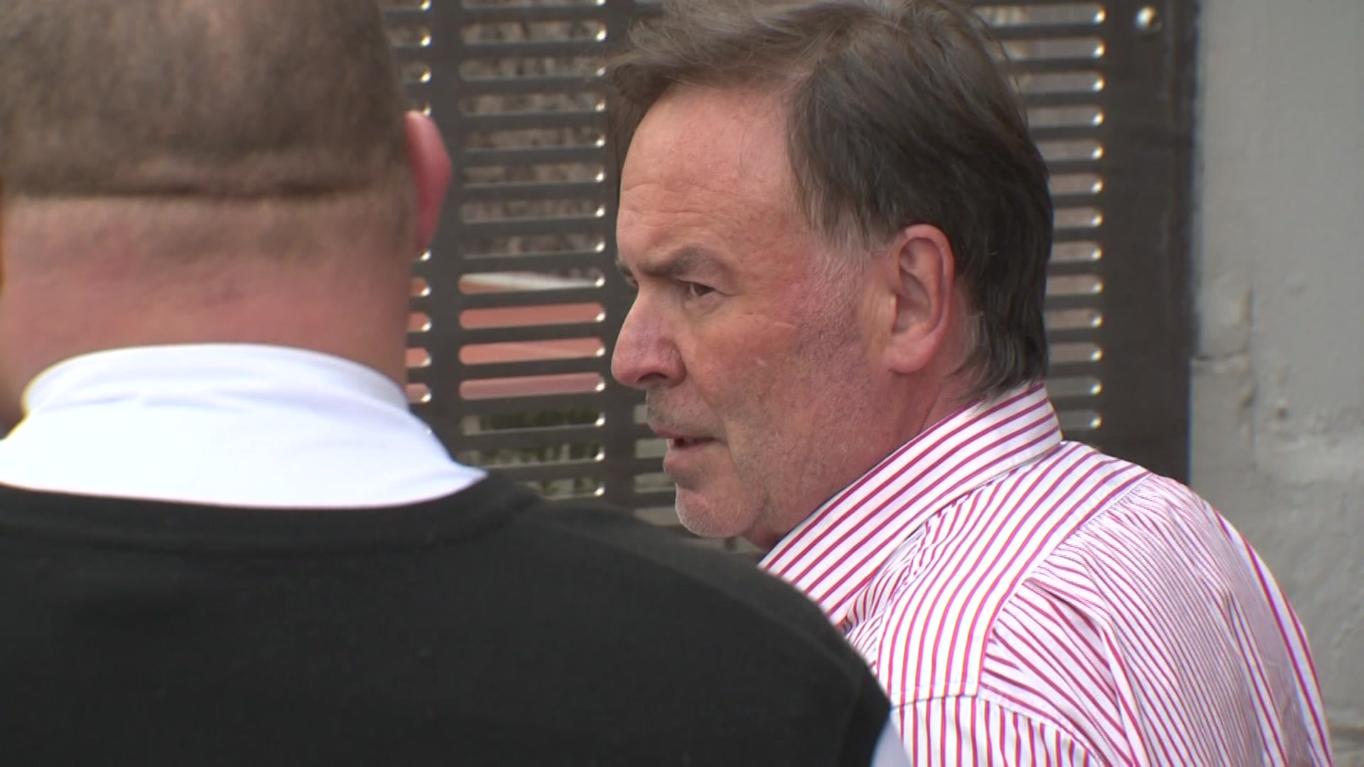 A middle-aged man stands in profile with a security on his left. 