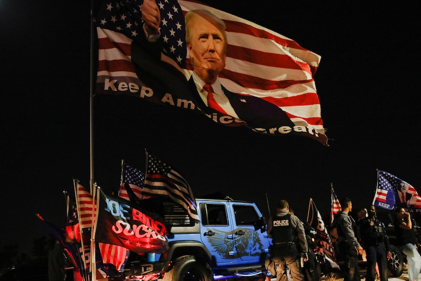 Men carrying US flags and flags bearing Donald Trump's face stand next to a ute at night 
