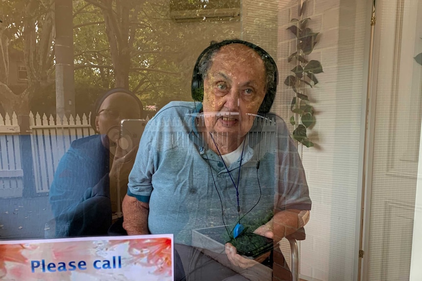 Older man wearing headphones holding phone looks through glass window where reflection of woman taking photo is visible