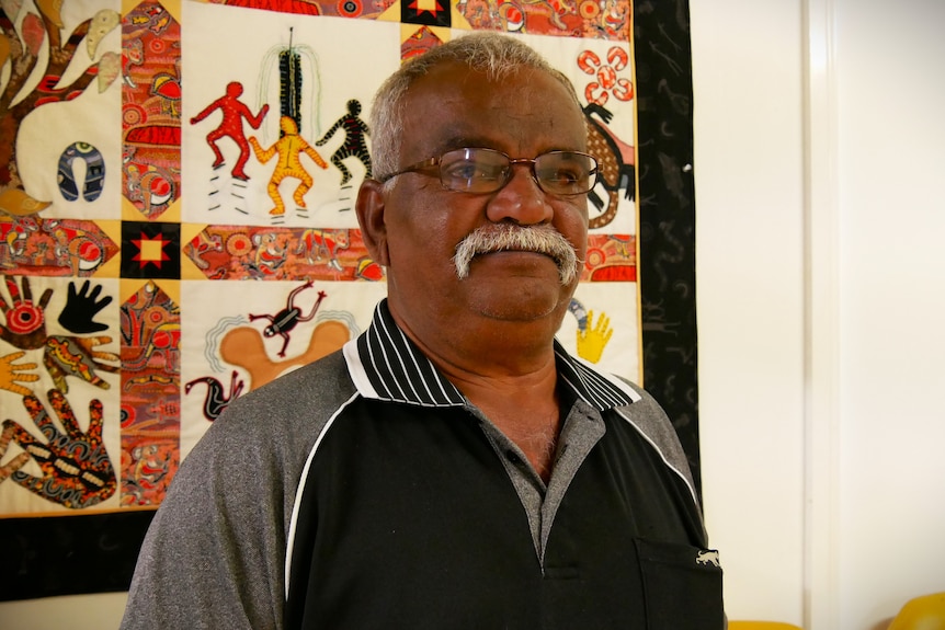 Older Aboriginal man with a white mustache and glasses, standing in front of an Aboriginal artwork