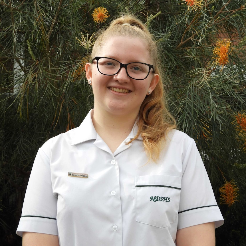 Girl in white school shirt and glasses smiles in front of native tree