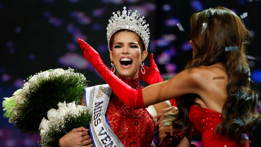 A woman wearing long red gloves places a large crown on the head of a smiling woman wearing a sash and clutching giant bouquet 