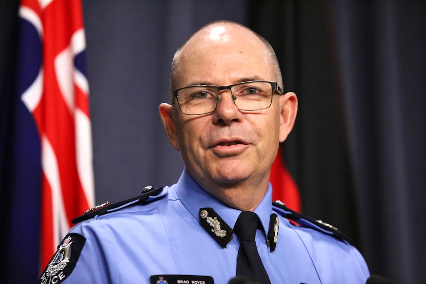 A man wearing glasses and a police uniform standing behind a flag and curtain