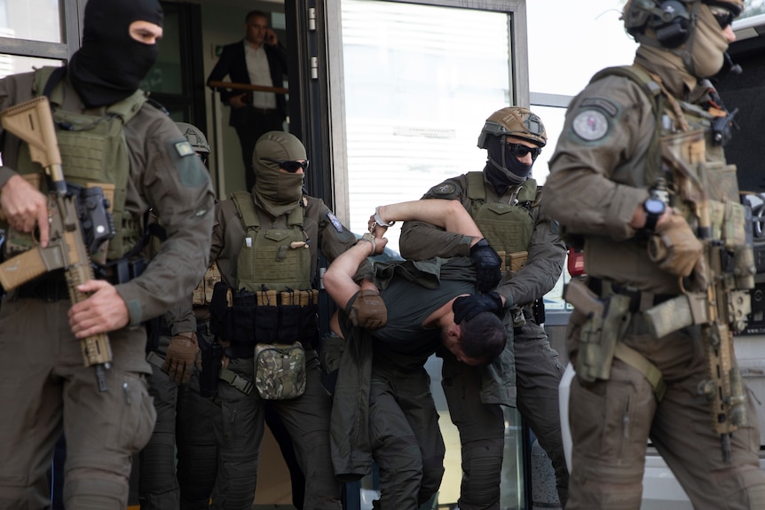 Armed police dressed in army-style fatigues and body armour manhandle a man in handcuffs.