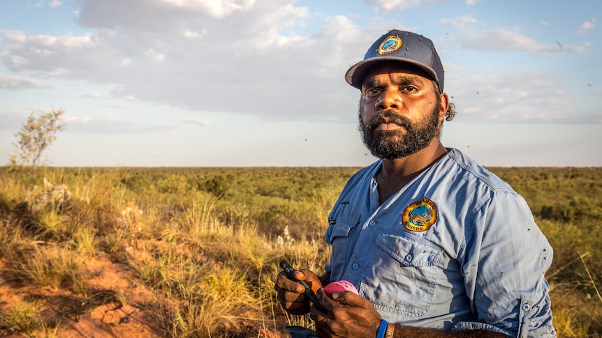 An aboriginal man stands in a uniform on top of a sand dune in the Australian desert.