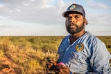 An Indigenous man wearing a ranger shirt and cap stands in an open grassland holding a roll of fluoro market tape.