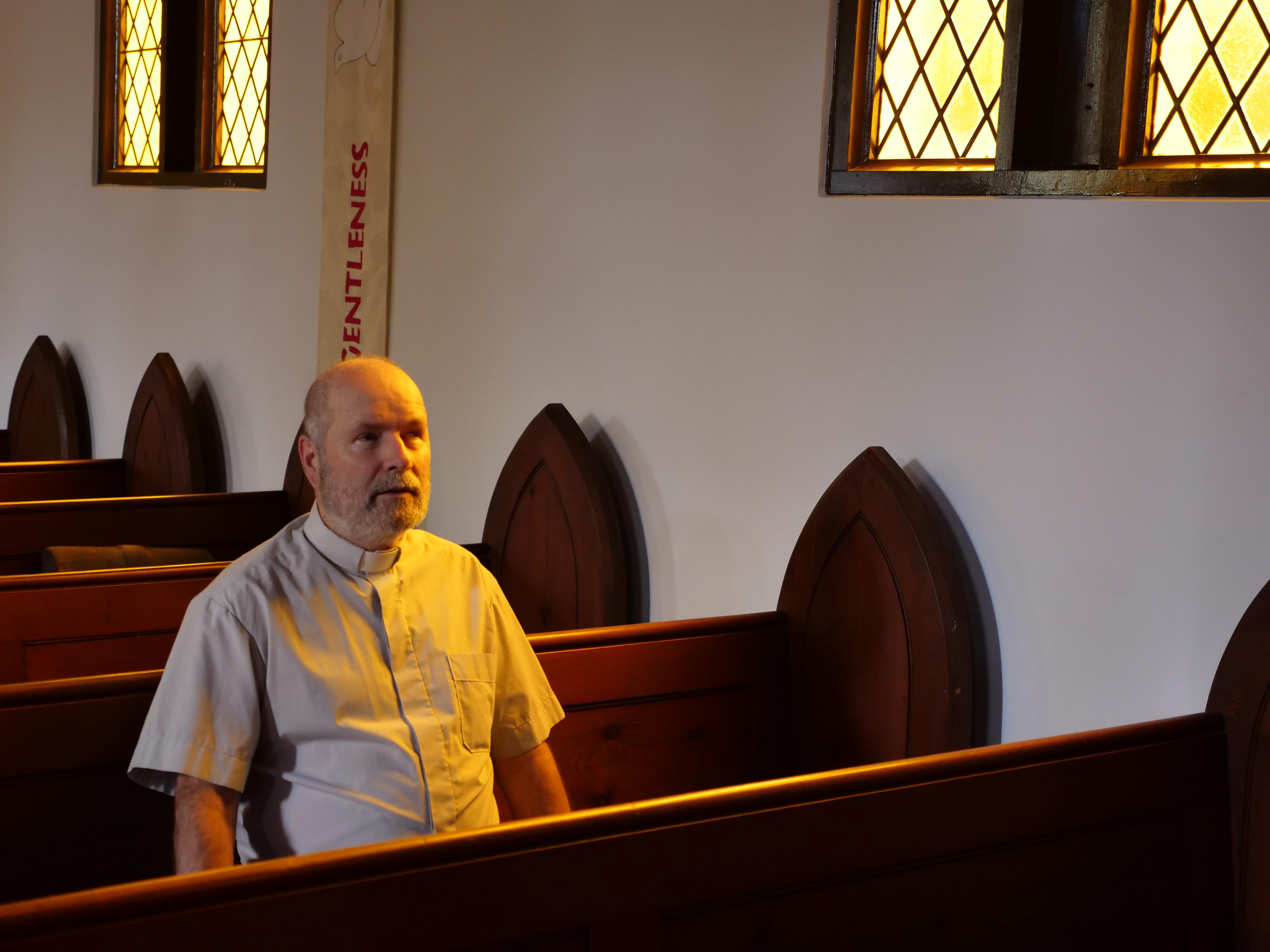 Frank se sienta en una iglesia, iluminada por una vidriera, claustros de madera.