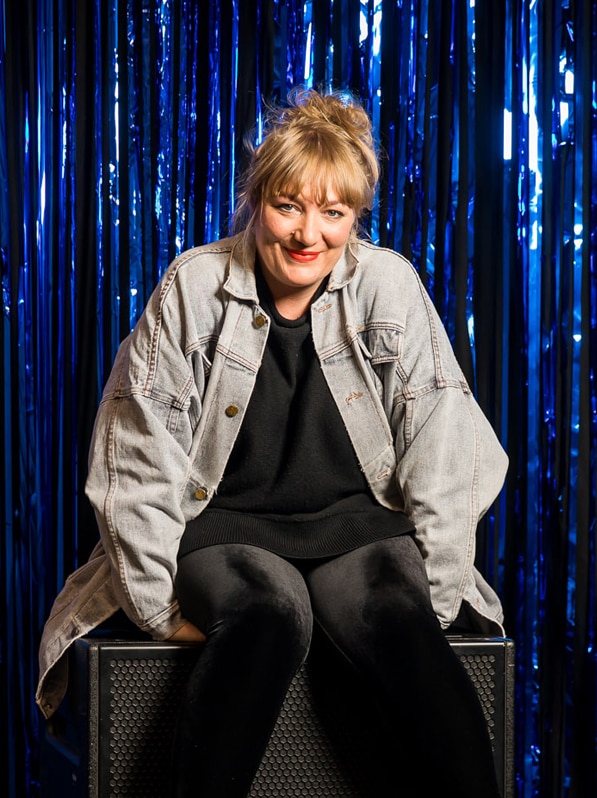 Paige Rattray sits on a black box in front of a blue foil tinsel curtain.