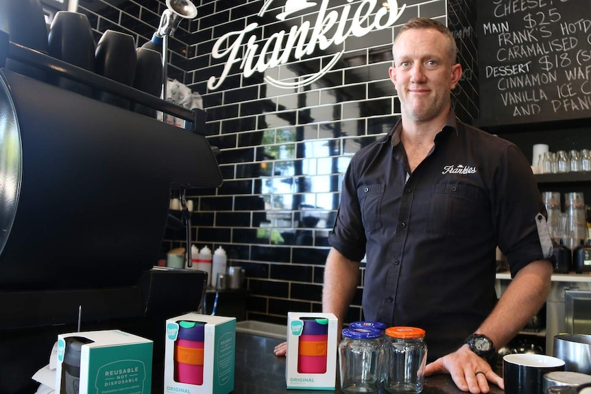 A man wearing a black shirt stands near a coffee machine, with jars and Keep Cups sitting on the bench.