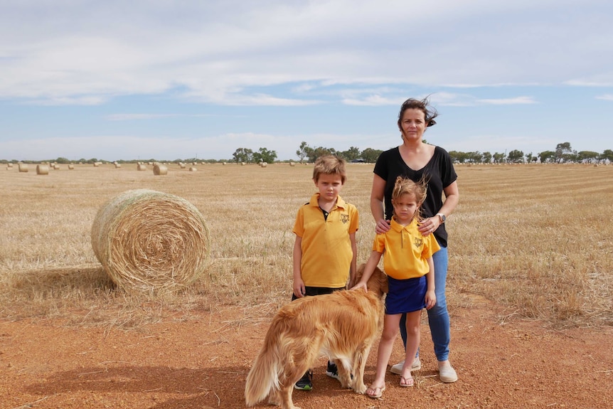 A woman stands in a golden coloured field with her 2 children and dog. There are hay bales dotted across the field.