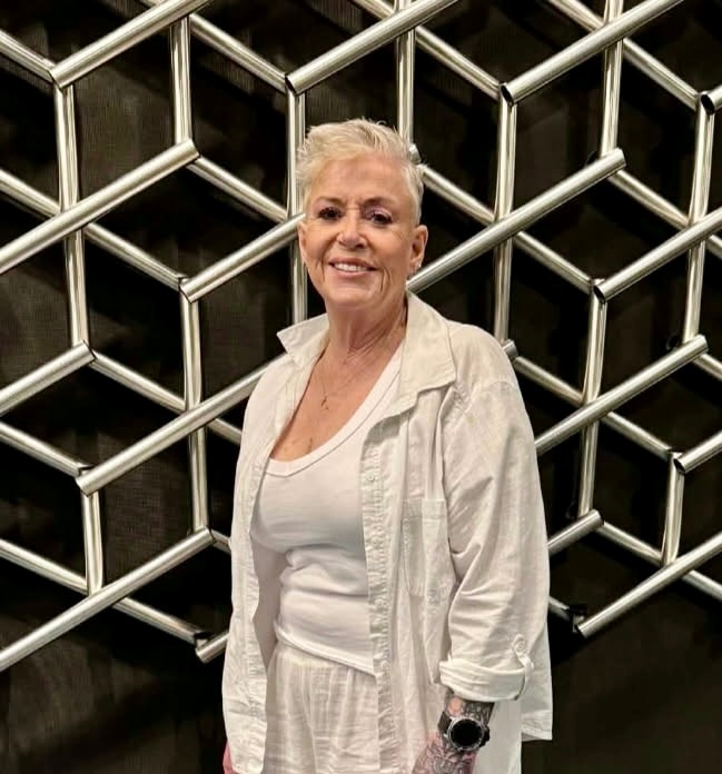 A woman with short grey hair stands in white linen next to a wall.
