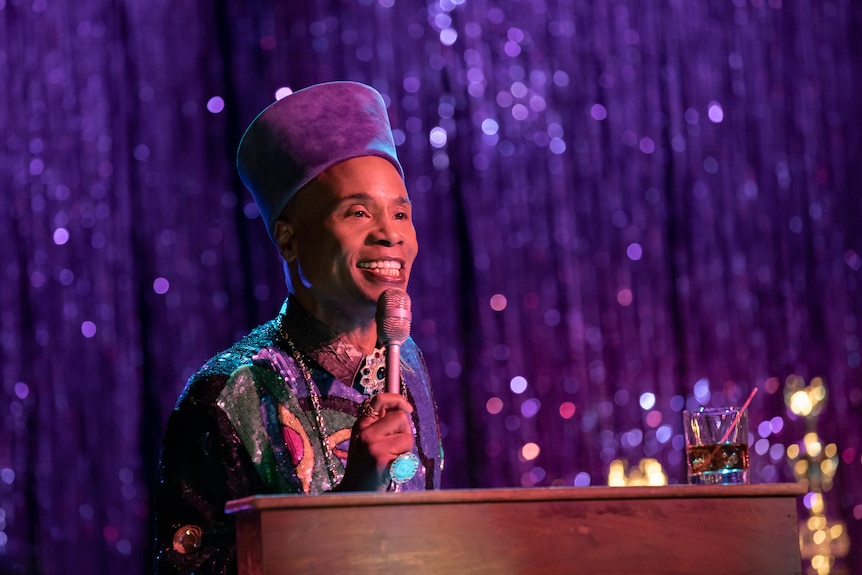 A black man with a purple hat speaking into a microphone, purple tinsel curtain in the background, in TV series Pose