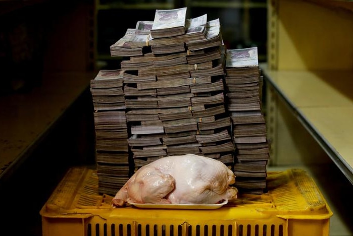 A raw chicken is seen in front of a tall stack of bolivar notes, both on top of a yellow crate with a dark backdrop.