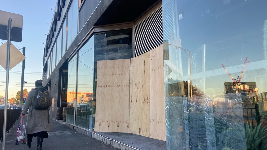 A glass shopfront facing a sidewalk has it's doors covered with wood panels.