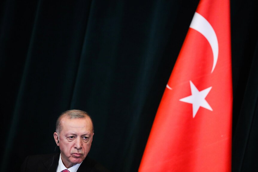 Recep Tayyip Erdogan looks down in front of a black background while standing next to the Turkish flag