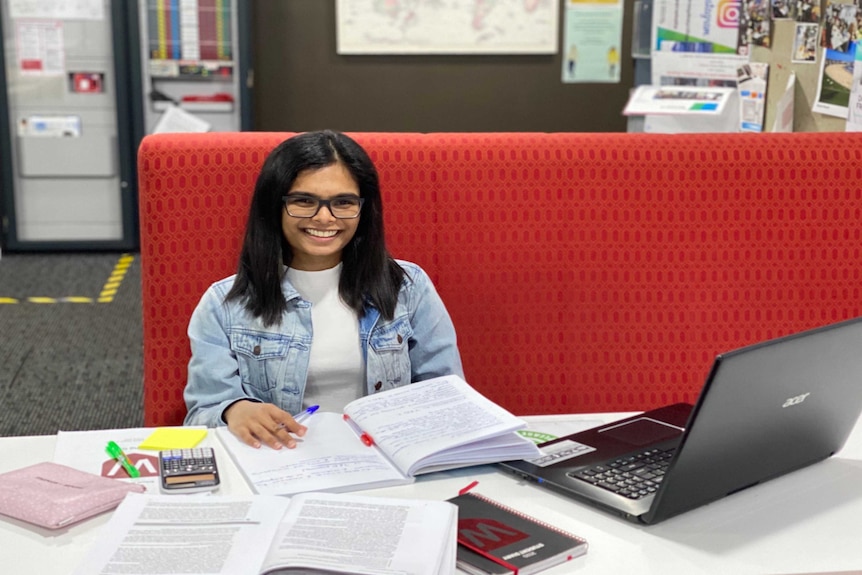 A woman with black hair and glasses studies at a desk, with books and her laptop open.