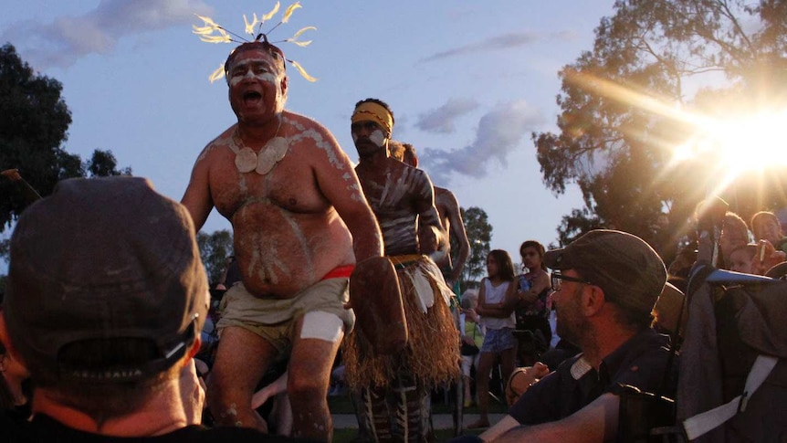An aboriginal man sings amongst a crowd, in white body paint and a feather headdress.