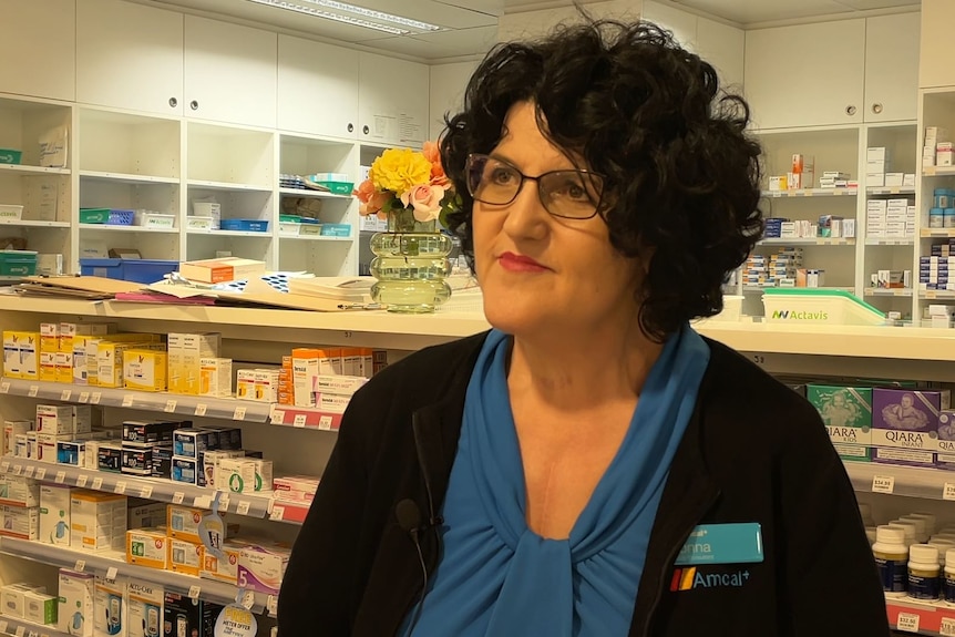 A lady with big black hair looks past the camera, stansd in a pharmacy, a name tag on her black sweater.