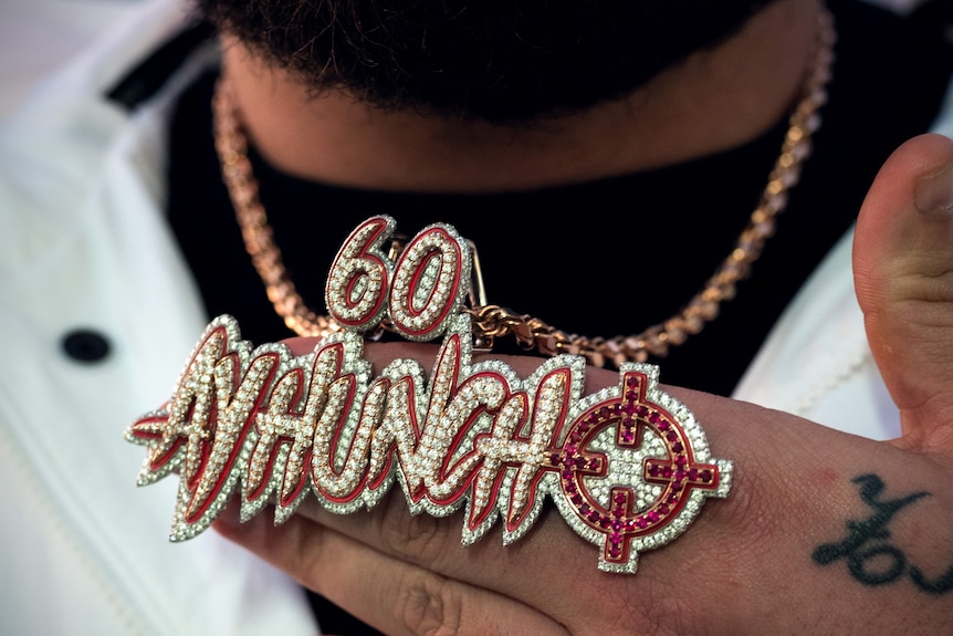 A man's hand holds up a diamond studded chain that says '60 Ay Huncho'. The 'O' is styled like a target sight symbol.