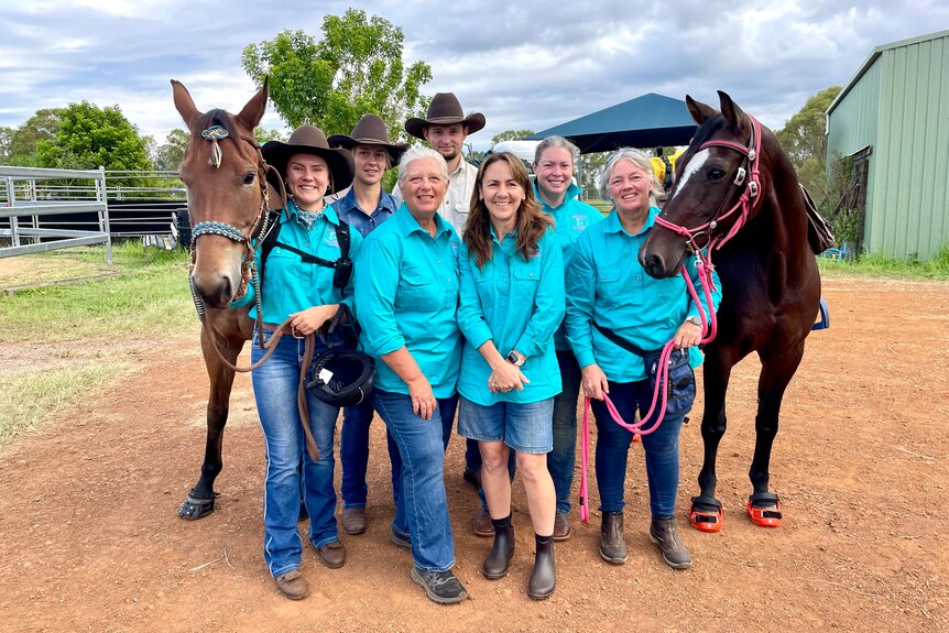 Men and women in aqua green shirts smile next to two horses.